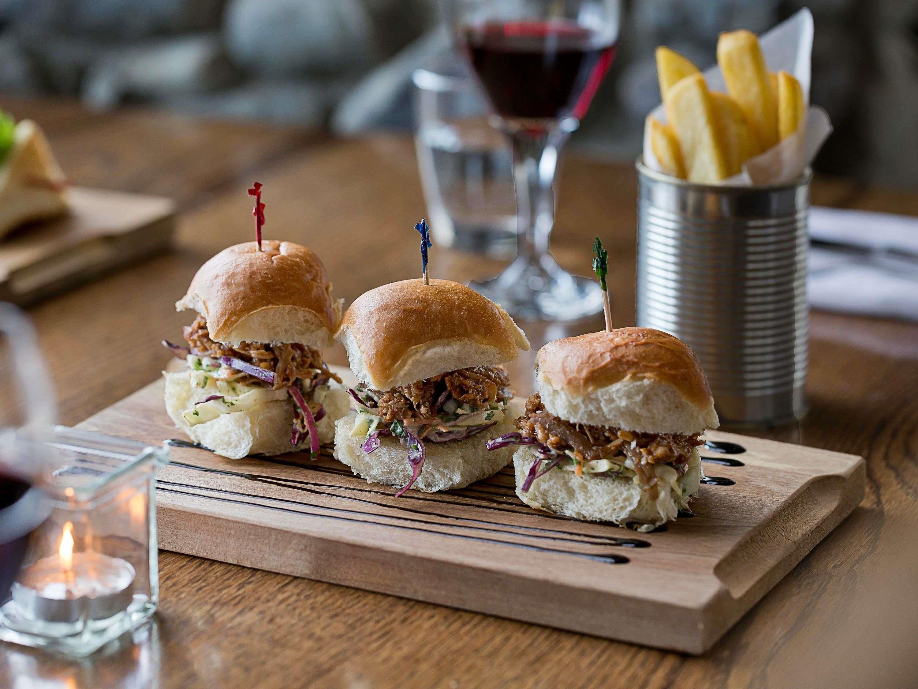Tres sándwiches sobre una tabla de madera, con una copa de vino tinto y un recipiente con papas fritas al fondo.