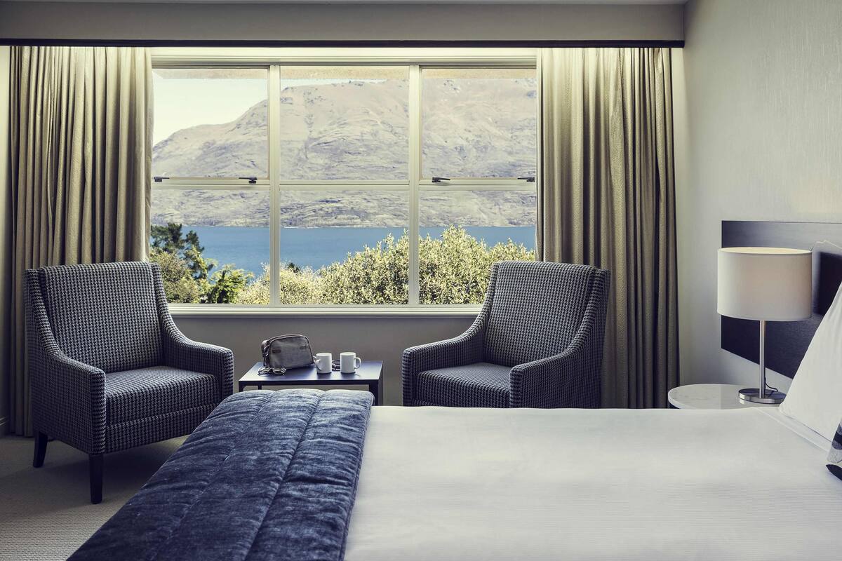 Ein Hotelzimmer mit einem großen Bett, zwei Sesseln, einem kleinen Tisch mit Lampe und Blick auf einen See und Berge durch ein großes Fenster.