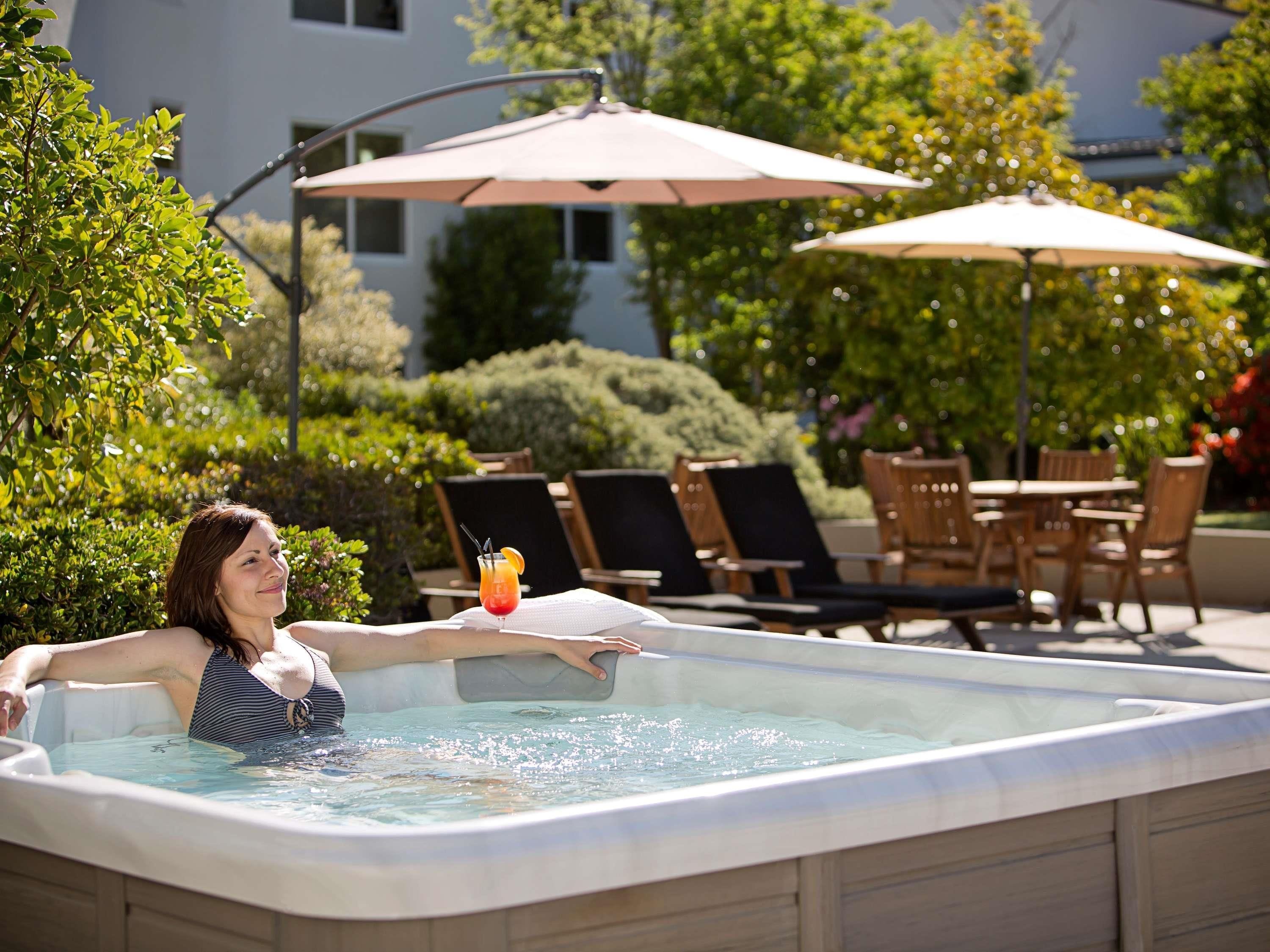 A person in a hot tub with a drink, surrounded by outdoor furniture and umbrellas.