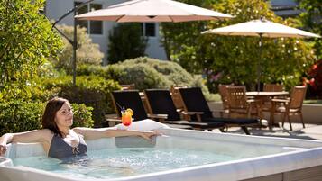 A person in a hot tub with a drink, surrounded by outdoor furniture and umbrellas.