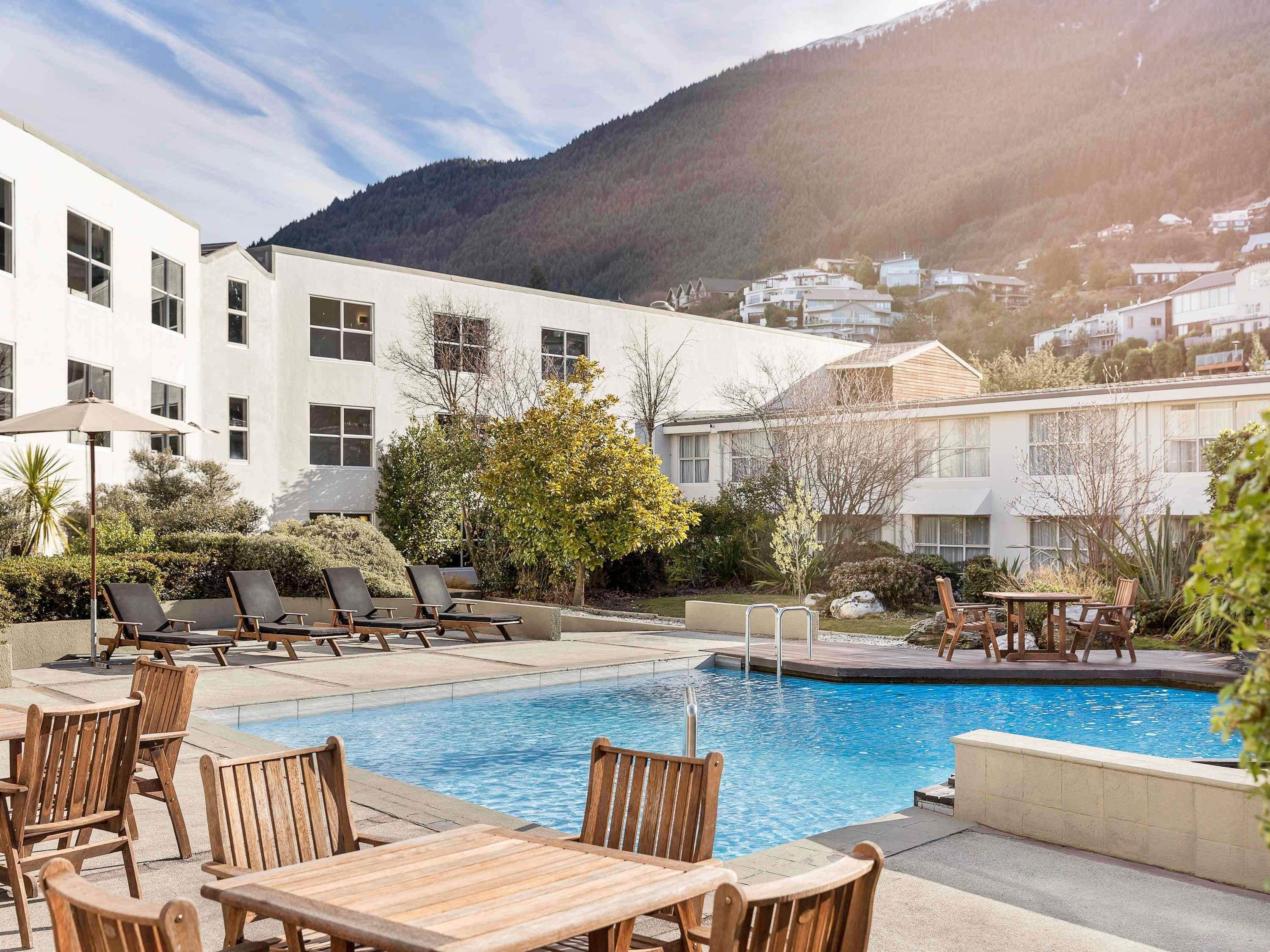 A pool area with lounge chairs and tables, surrounded by a building with large windows and a mountain backdrop.