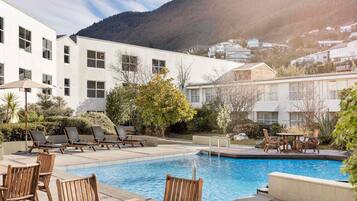 A pool area with lounge chairs and tables, surrounded by a building with large windows and a mountain backdrop.