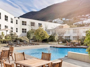 A pool area with lounge chairs and tables, surrounded by a building with large windows and a mountain backdrop. - Mövenpick Queenstown (Queenstown)
