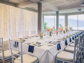 Uma sala de banquetes com uma longa mesa posta para um evento formal, grandes janelas com vista para as montanhas e um teto com iluminação embutida.