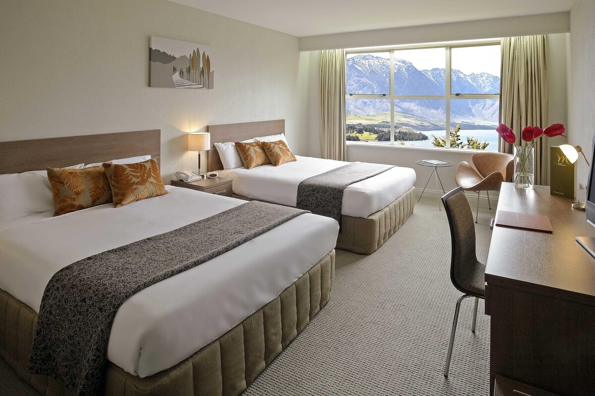 A hotel room with two beds, a desk, a chair, a view of mountains, and a painting on the wall.