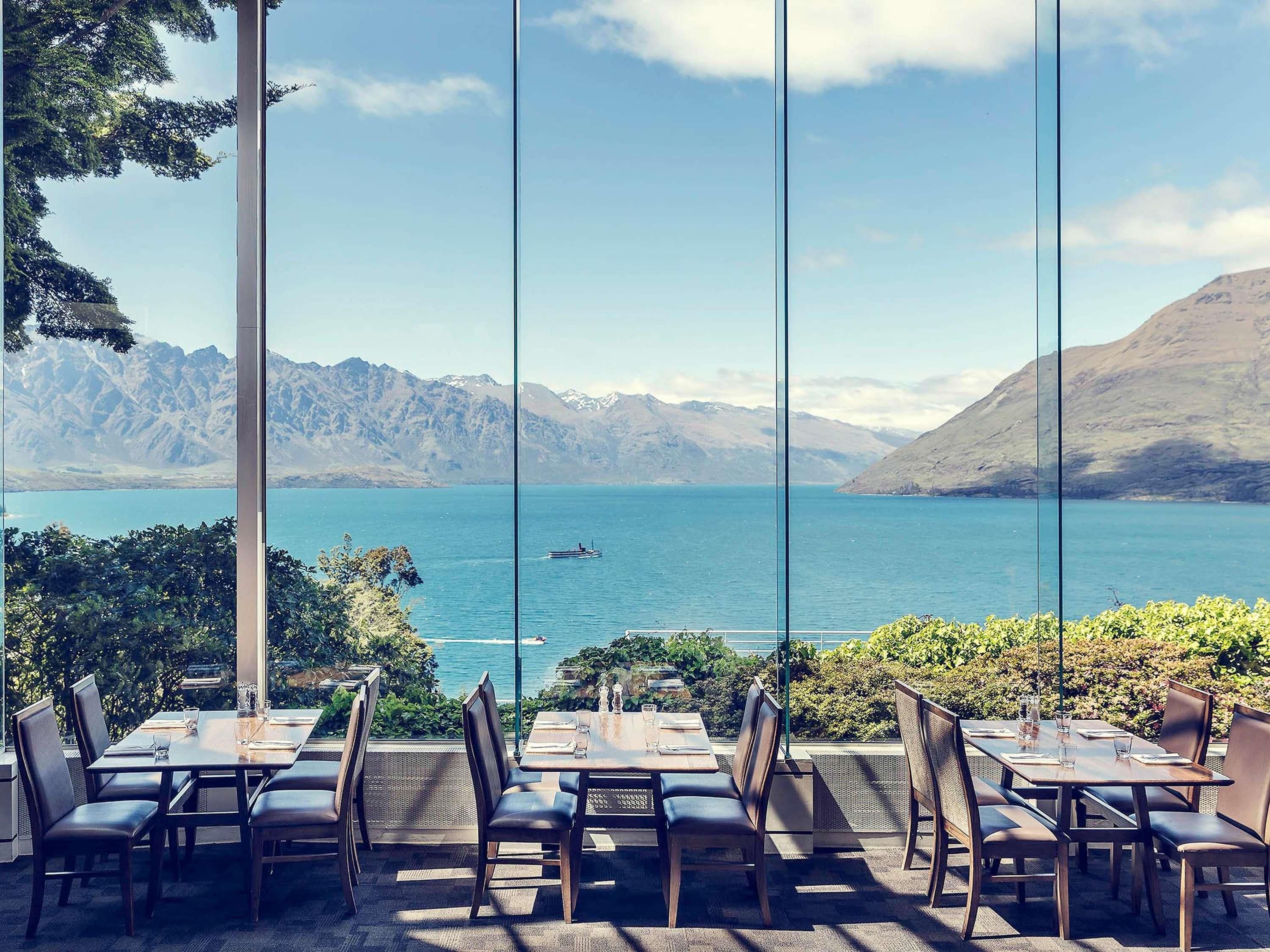An outdoor dining area with tables and chairs, overlooking a lake with mountains in the background.