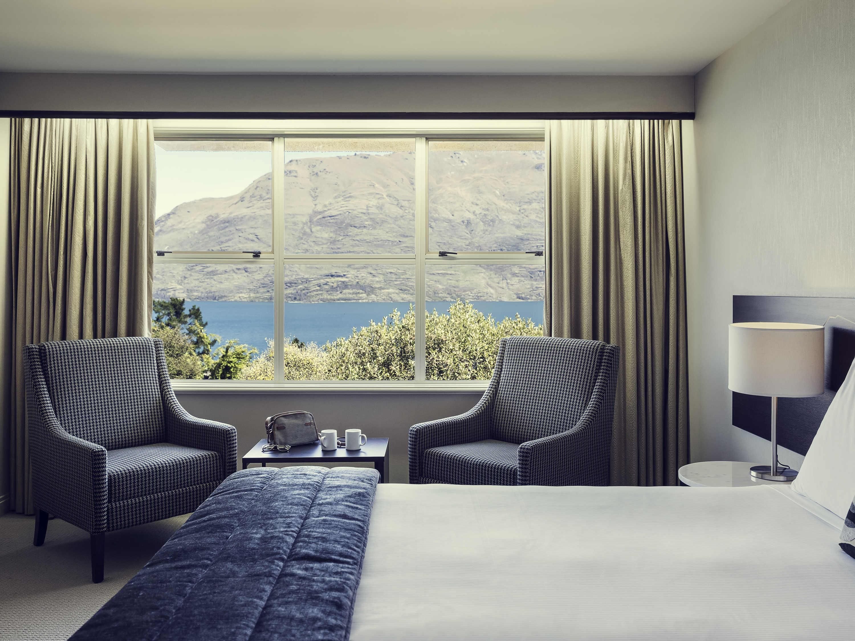 A hotel room with a large bed, two armchairs, a small table with a lamp, and a view of a lake and mountains through a large window.