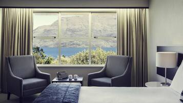 A hotel room with a large bed, two armchairs, a small table with a lamp, and a view of a lake and mountains through a large window.