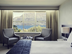 A hotel room with a large bed, two armchairs, a small table with a lamp, and a view of a lake and mountains through a large window. - Mövenpick Queenstown (Queenstown)