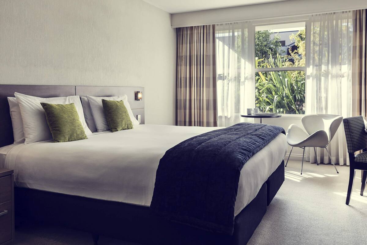 Ein Hotelzimmer mit einem großen Bett, einem Schreibtisch, einem Sessel und einem Fenster mit Vorhängen.
