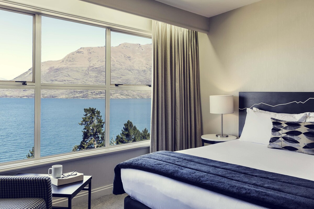 A hotel room with a large window, a bed, a bedside table, a lamp, and a view of a body of water and mountains.
