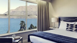 A hotel room with a large window, a bed, a bedside table, a lamp, and a view of a body of water and mountains. - Mercure Queenstown Resort (Queenstown)