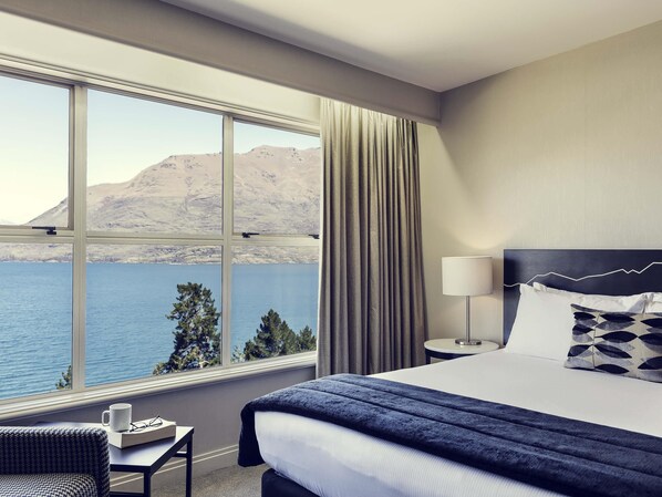 Ein Hotelzimmer mit großem Fenster, einem Bett, einem Nachttisch, einer Lampe und Blick auf ein Gewässer und Berge.