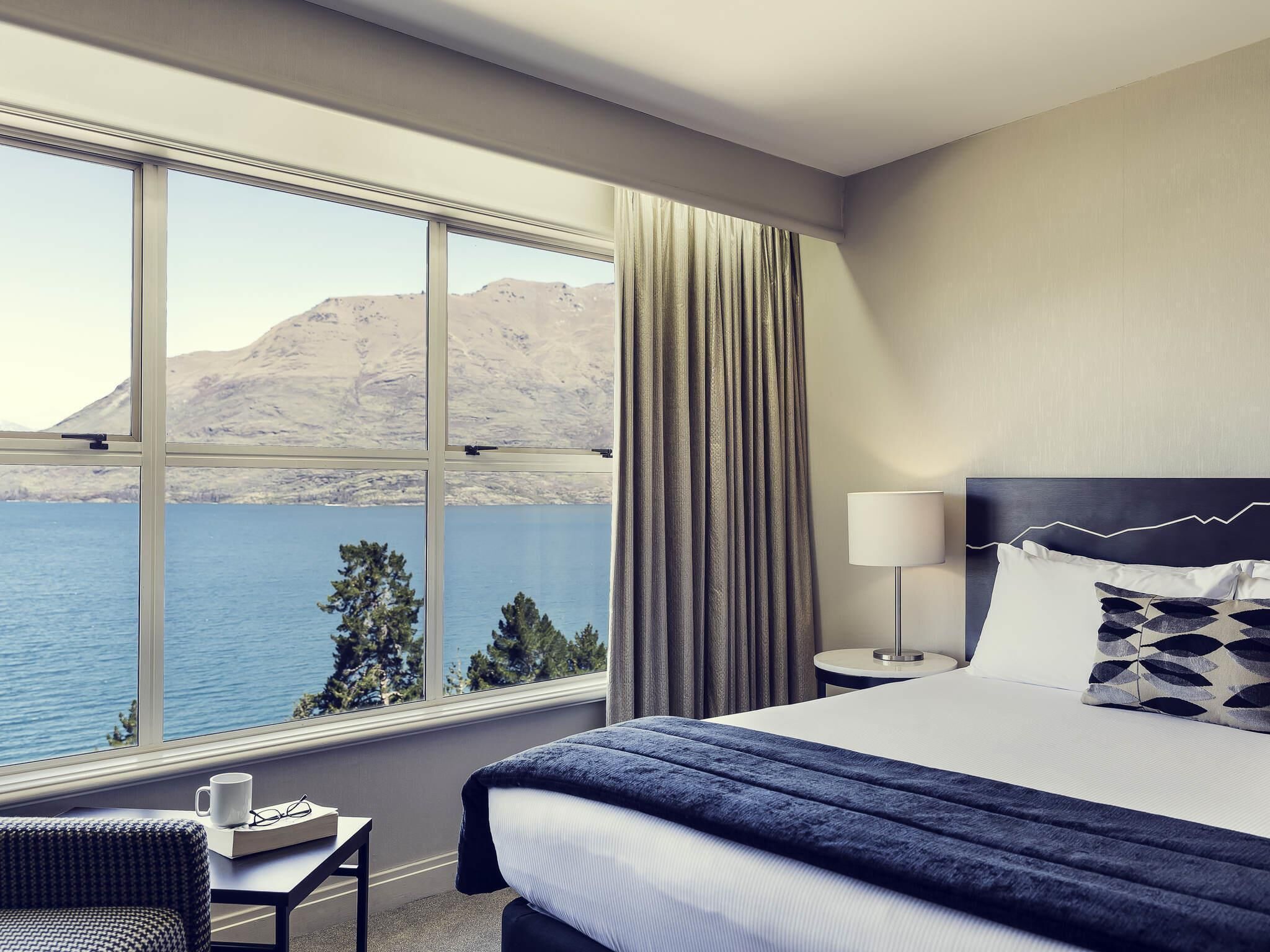 A hotel room with a large window, a bed, a bedside table, a lamp, and a view of a lake and mountains.