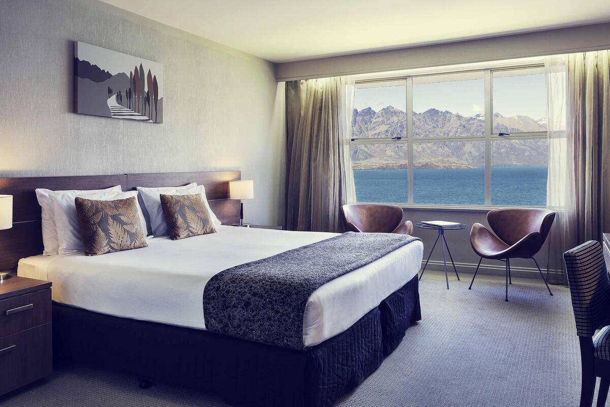 Ein Hotelzimmer mit einem großen Bett, zwei Sesseln, einem kleinen Tisch, einem Nachttisch und einem Fenster mit Blick auf die Berge.