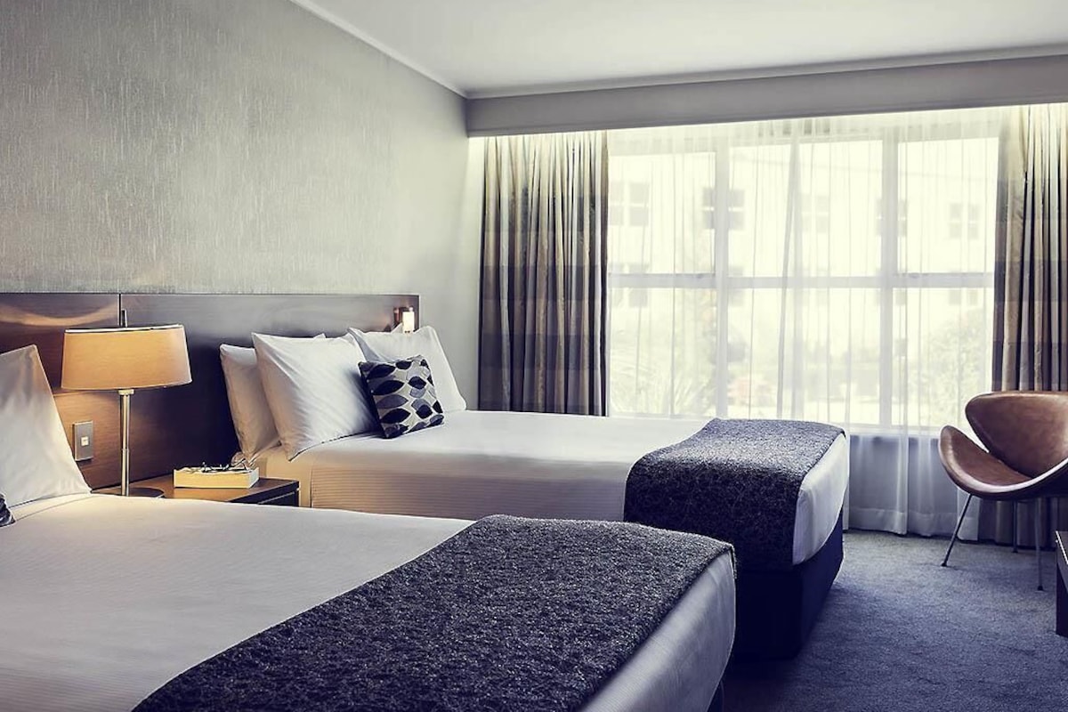 A hotel room with two beds, a chair, a nightstand, and a large window with curtains.