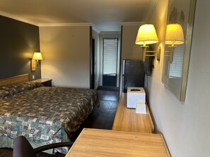 A hotel room with a large bed, a wooden desk, a chair, two wall-mounted lamps, and a refrigerator.