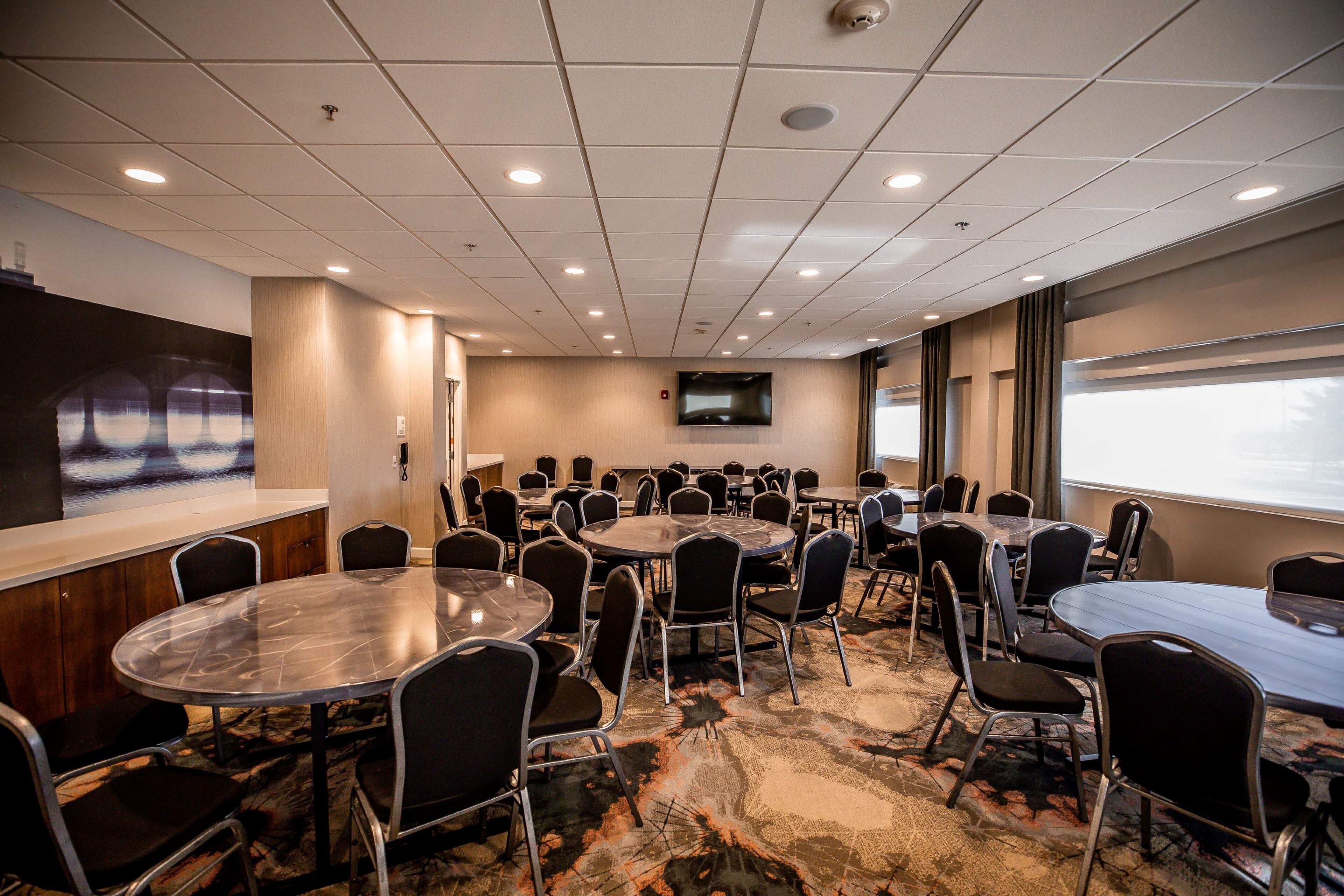 A conference room with multiple tables and chairs, a mounted TV, and a large window with a view.