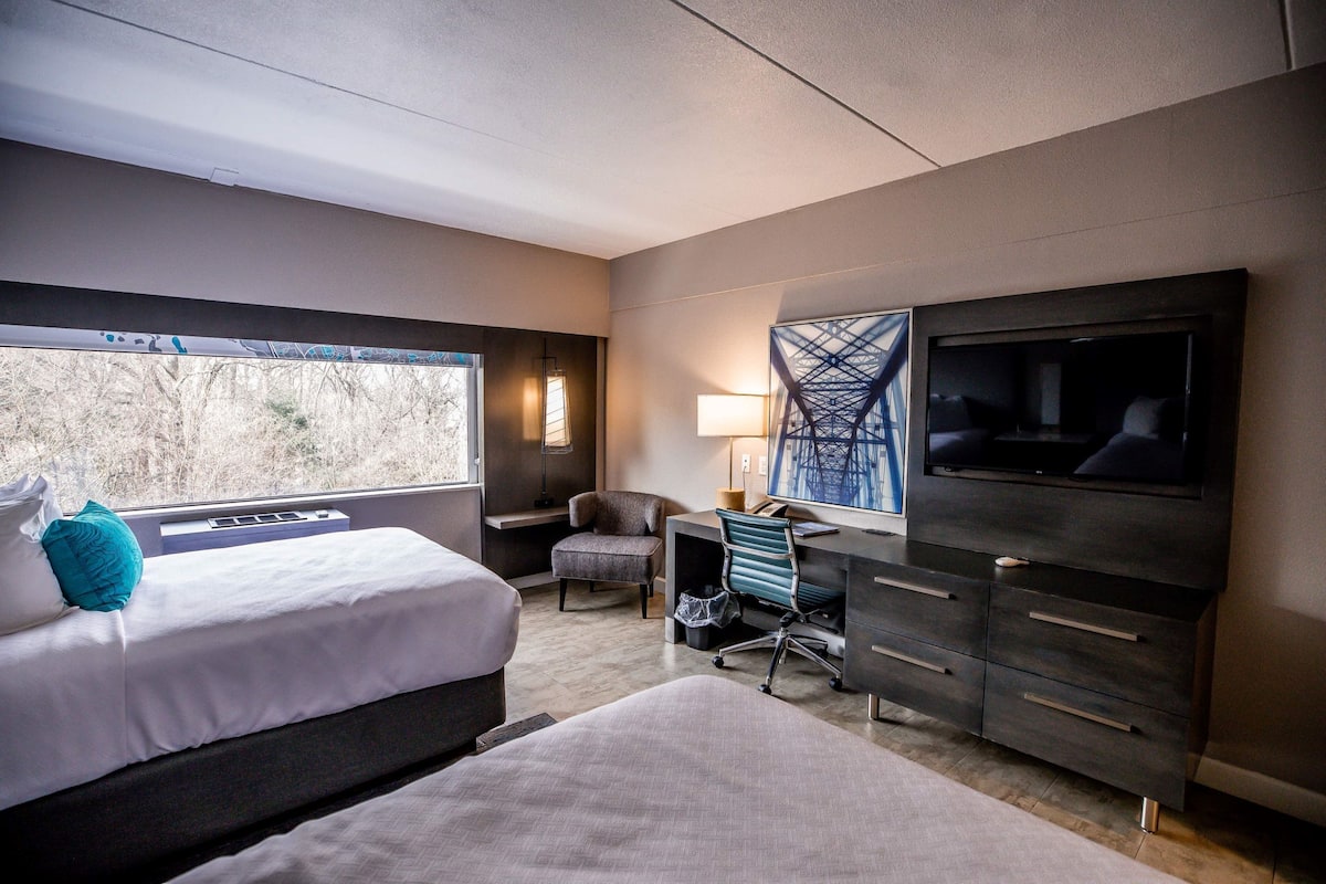 A hotel room with a bed, a desk, a chair, a TV, and a window with a view of trees.