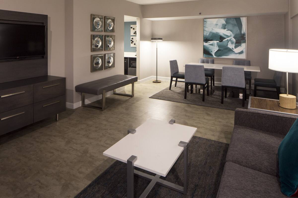 A modern living room with a flat-screen TV, a bench, a dining area with a table and chairs, and a large abstract painting on the wall.