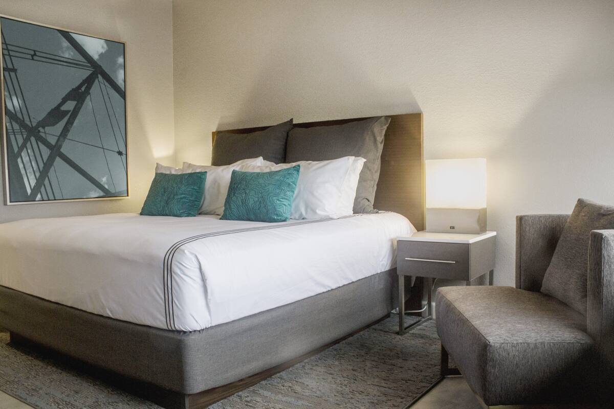 A modern hotel room with a large bed, a nightstand, a chair, and a wall-mounted lamp.