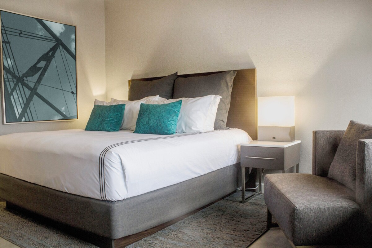 A modern hotel room with a large bed, a nightstand, a chair, and a wall-mounted lamp.