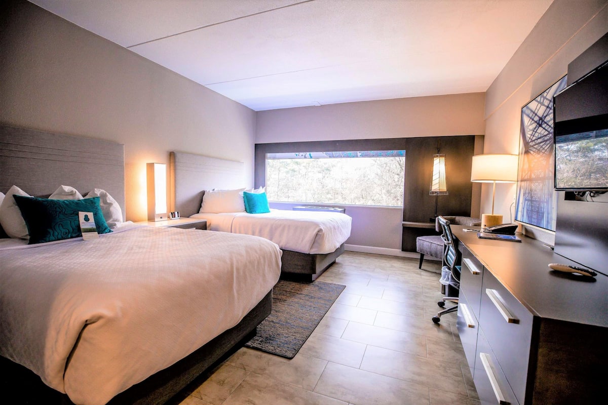 Habitación de hotel con dos camas, un escritorio con computadora, televisor y ventana con vistas.
