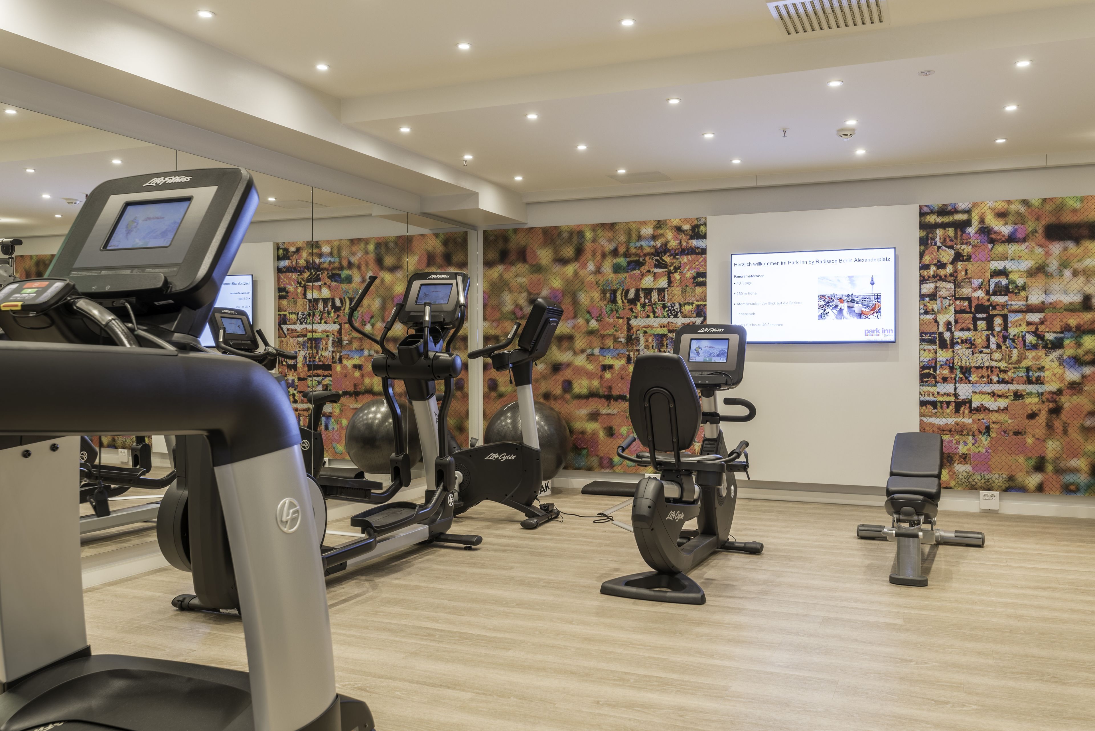 A modern gym with various exercise equipment, including treadmills and stationary bikes, and a digital screen displaying workout information.