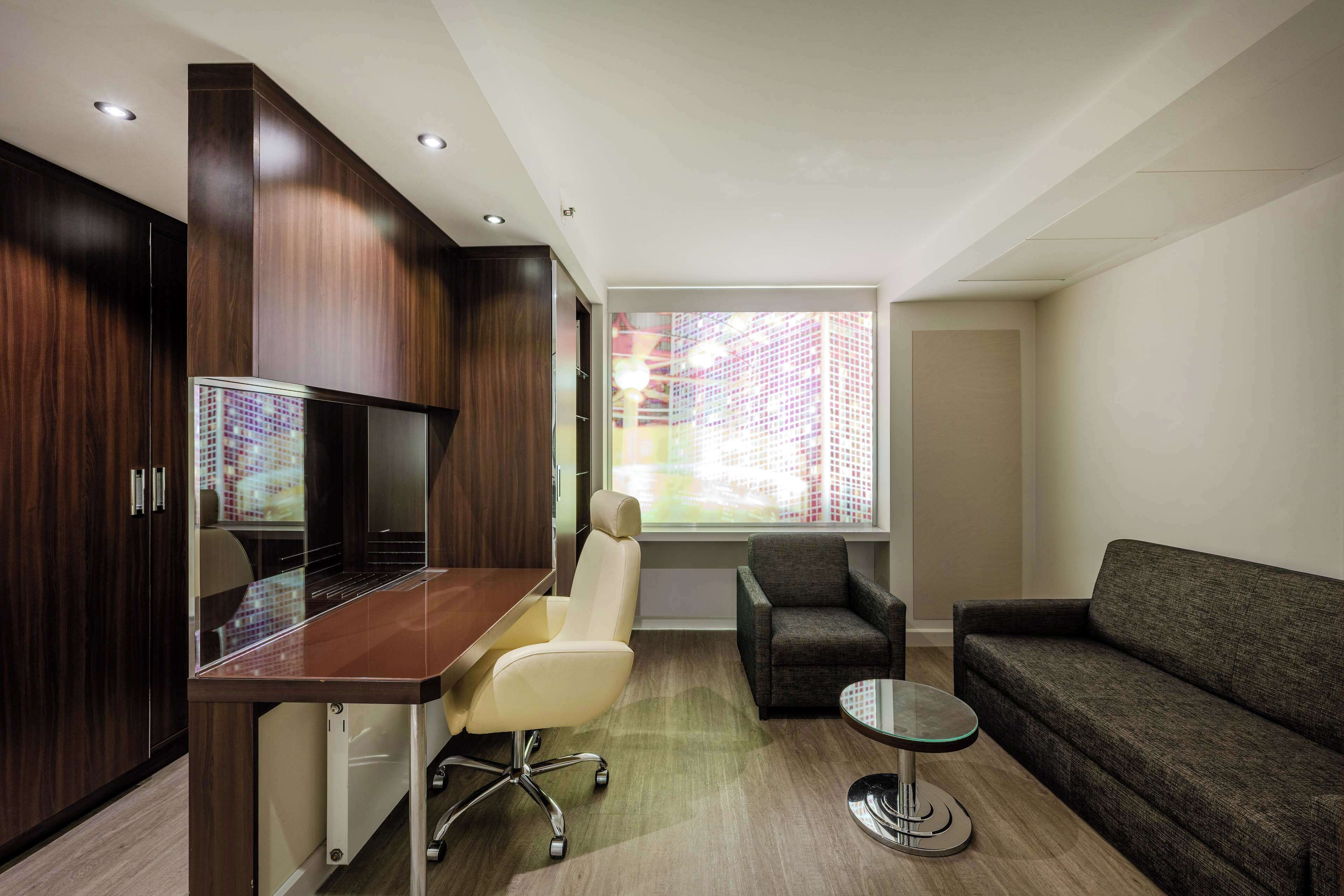 A modern living room with a wooden cabinet, a glass-top table, a leather chair, a grey sofa, and a small round table.