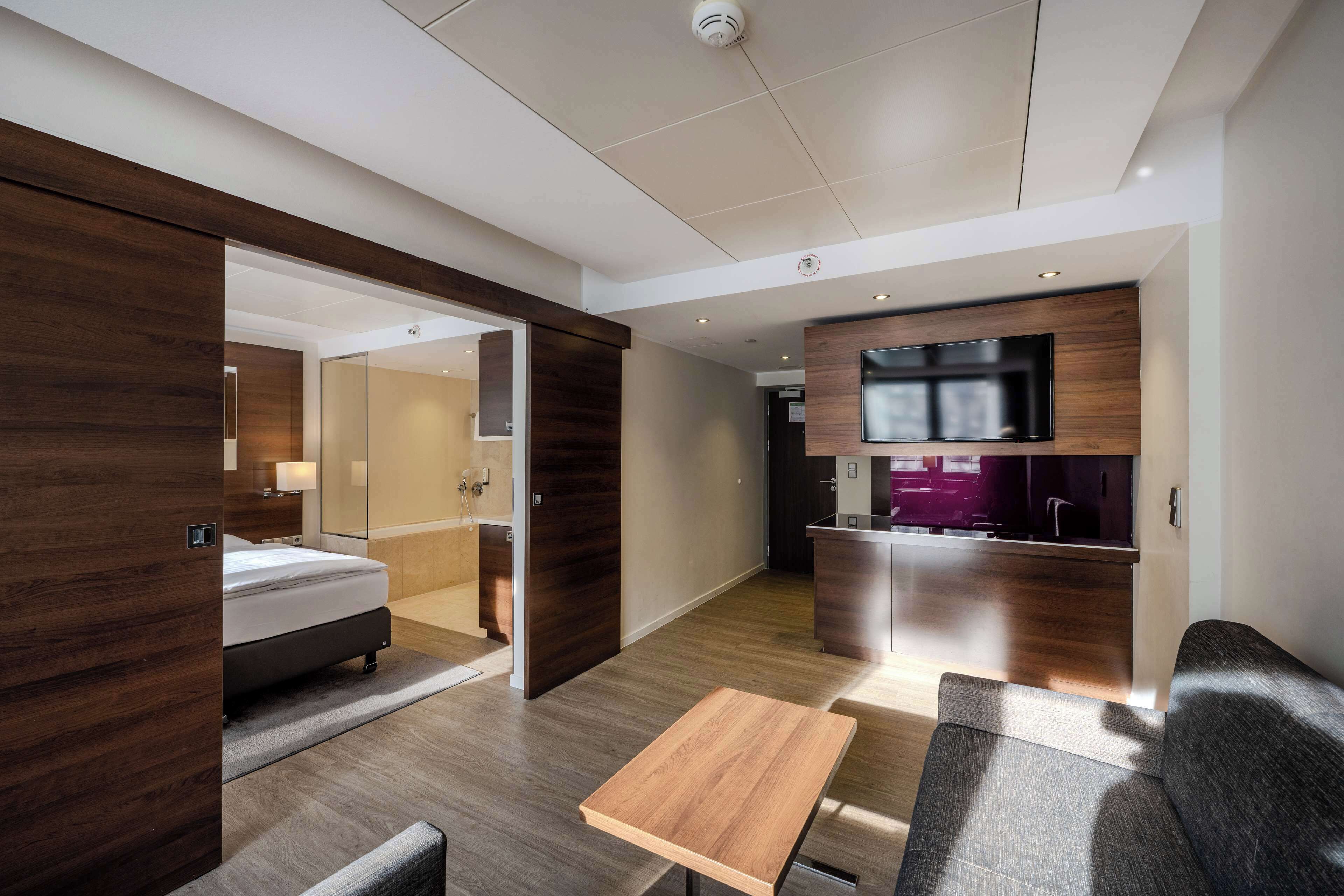 Una habitación de hotel moderna con suelo de madera, una cama, una pequeña mesa de madera, un sofá, un televisor montado en la pared y una zona de cocina con encimera morada.
