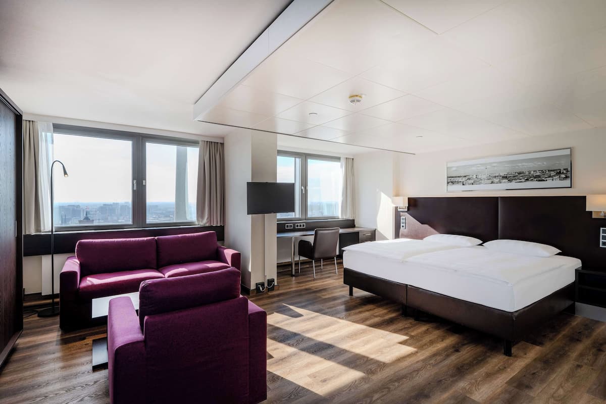 Una habitación de hotel moderna con una cama grande, un sofá, un escritorio y vistas a la ciudad a través de amplias ventanas.