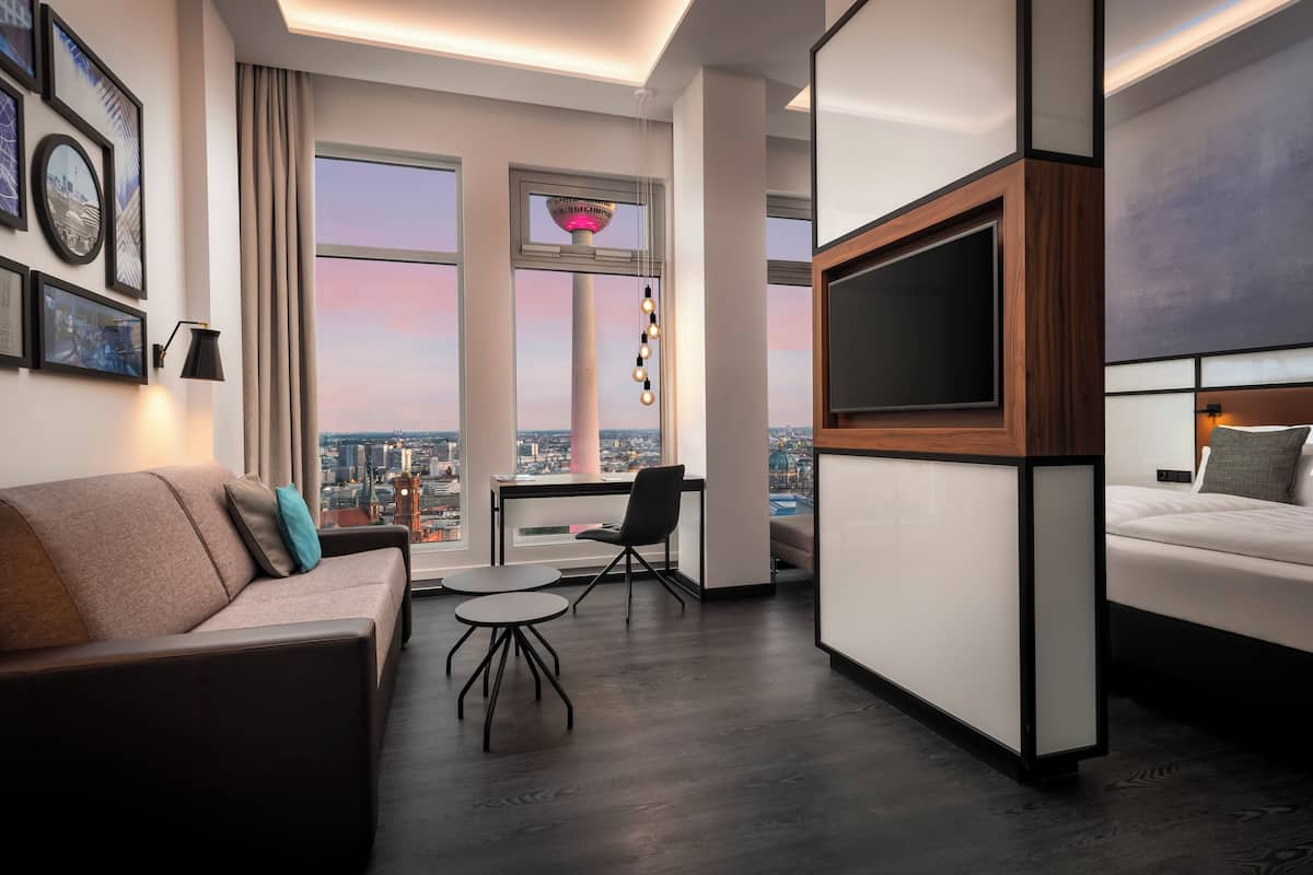Habitación de hotel moderna con un televisor grande, un sofá, un escritorio y vistas de la ciudad.