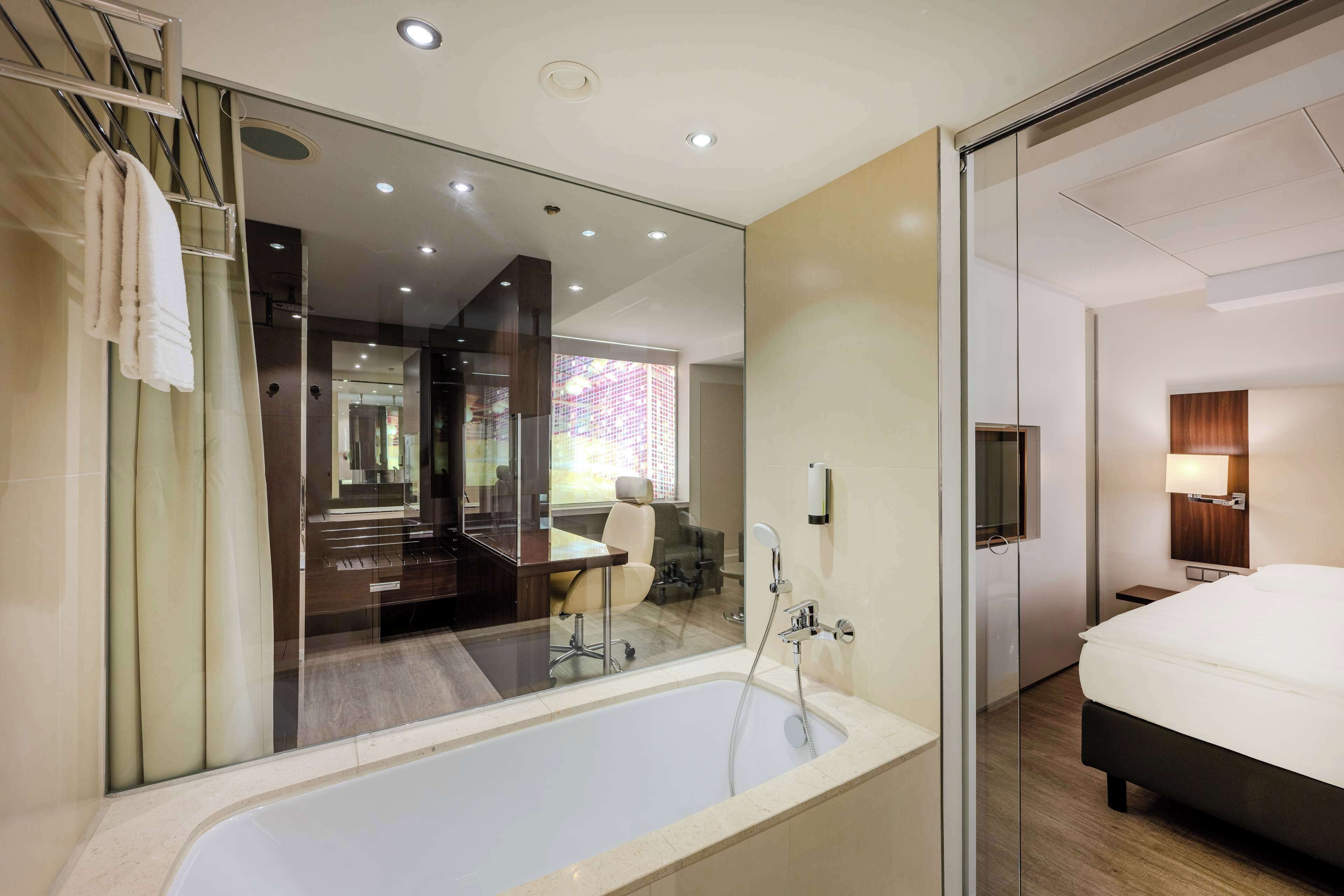 Una habitación de hotel moderna con una bañera grande, zona de ducha independiente, cama, escritorio y área de estar con vistas al exterior.