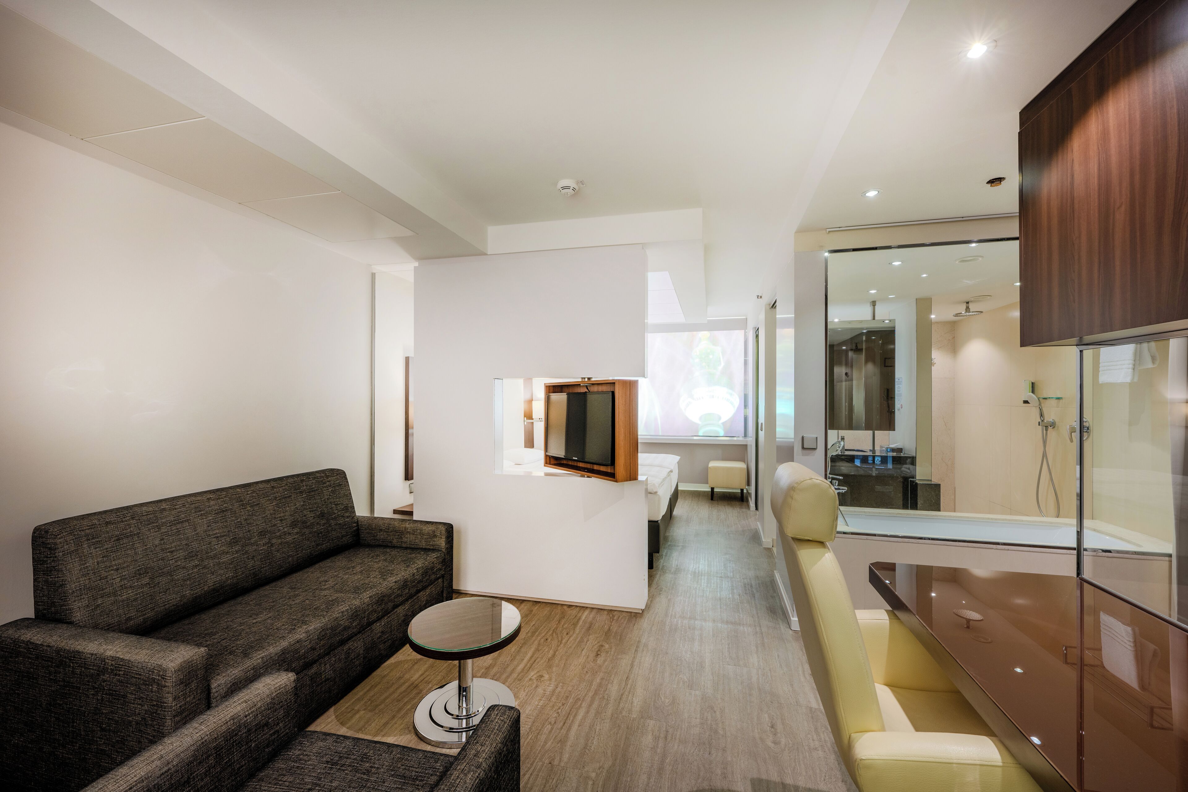 A modern hotel room with a sofa, a small round table, a bed, and a glass-enclosed shower area.