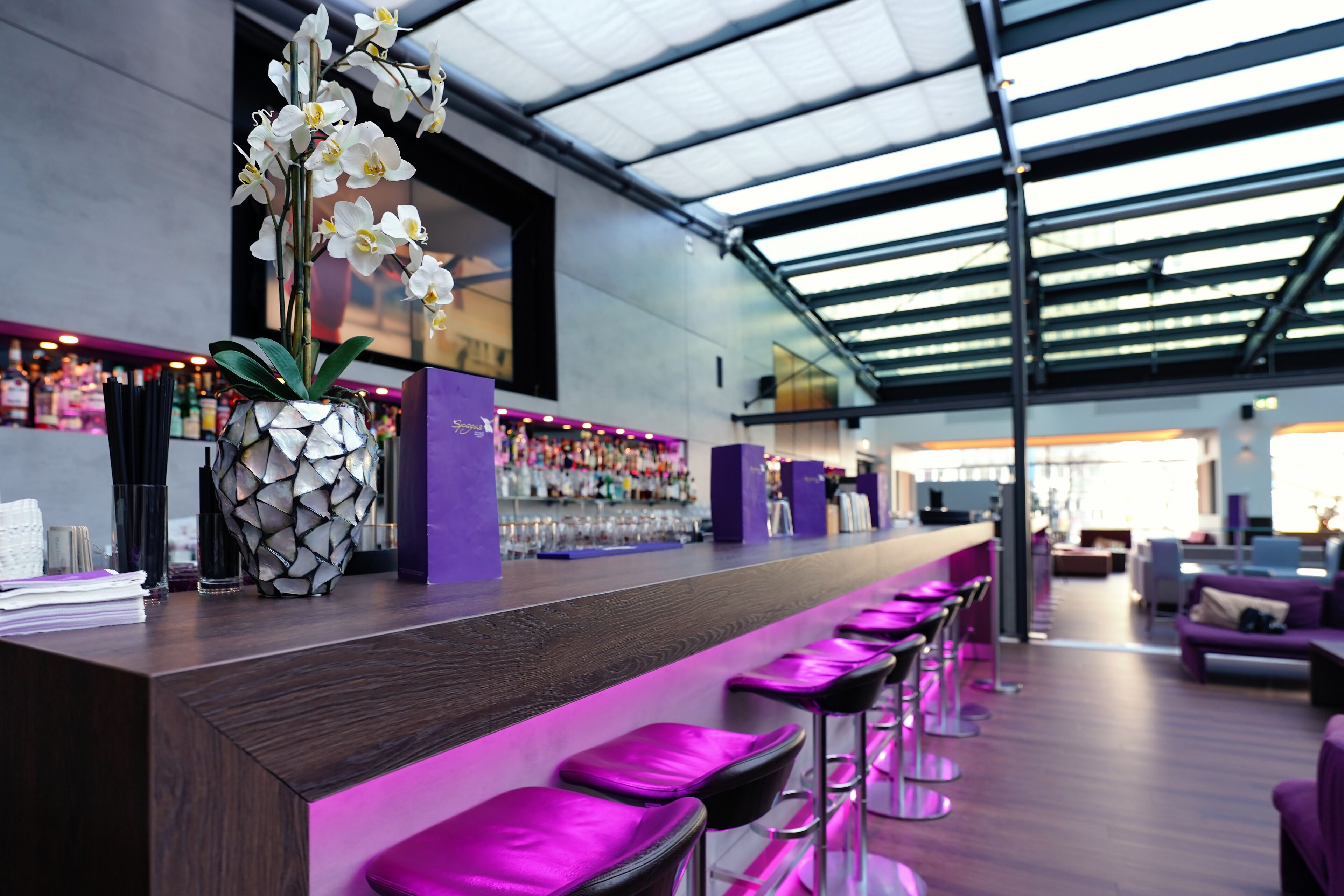 A modern bar area with a long counter, purple bar stools, and a decorative orchid arrangement.