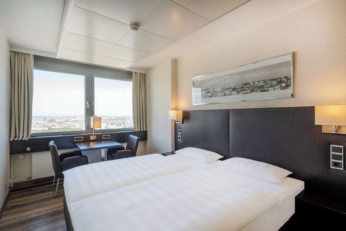 Habitación de hotel con dos camas, un escritorio y una silla. Cuenta con un ventanal con cortinas, un cuadro enmarcado en la pared y una lámpara fijada a la pared.