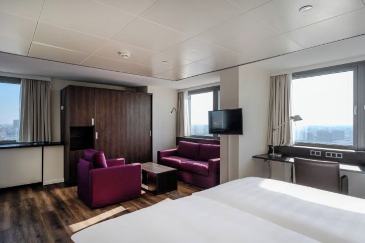 A modern hotel room with a large bed, two purple sofas, a small table, a TV, and a desk with a computer.