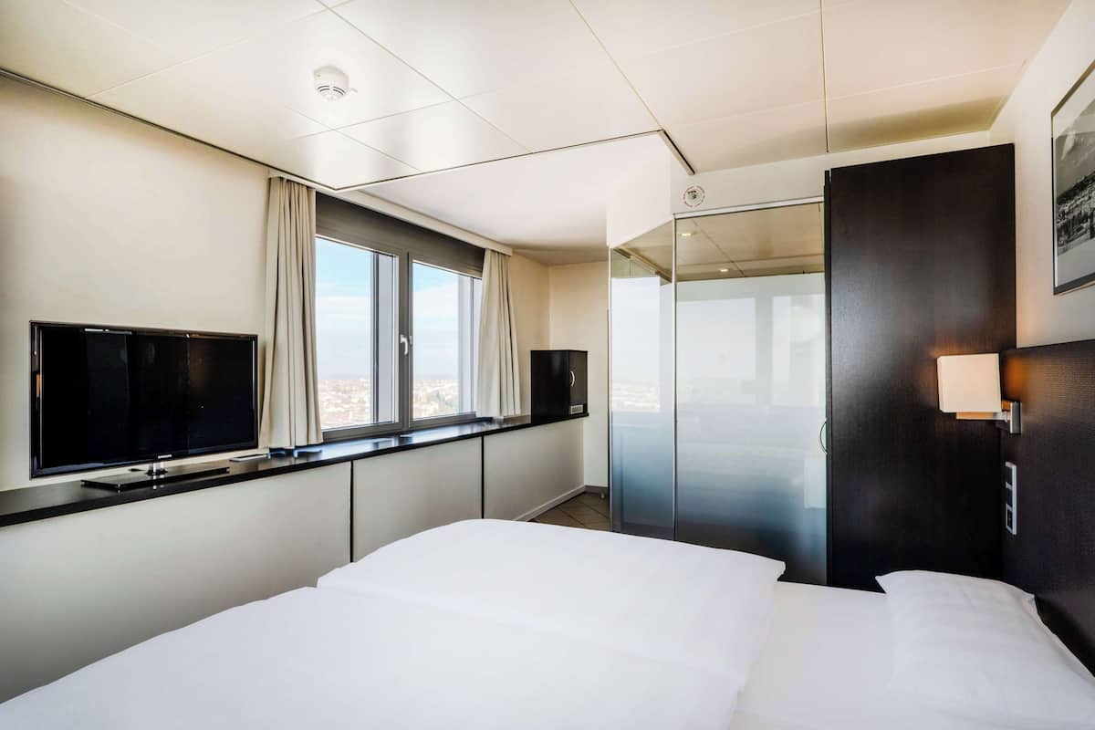 Una habitación de hotel moderna con una cama grande, un televisor de pantalla plana, una ventana con cortinas, una puerta de vidrio y una lámpara fijada a la pared.