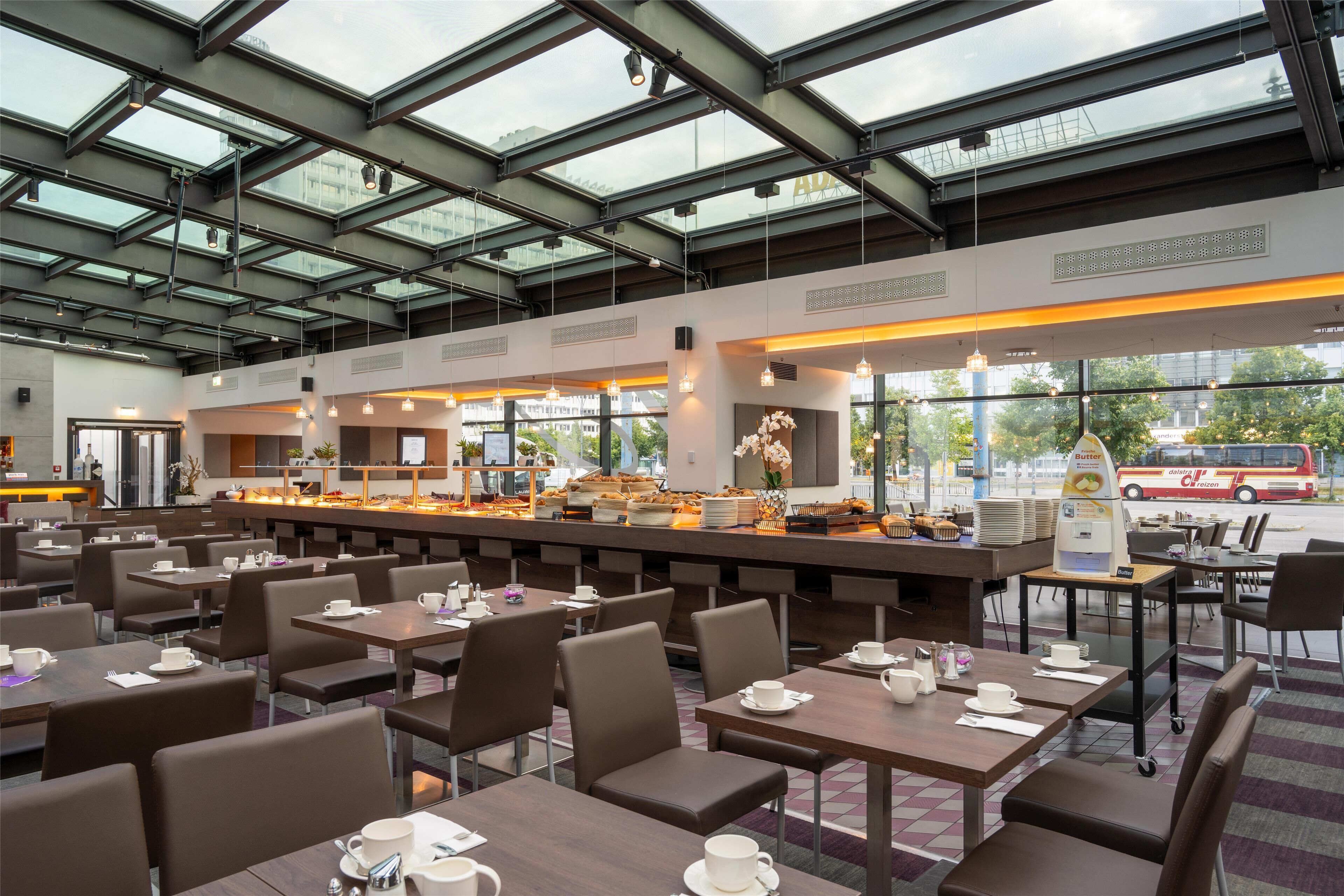 A modern cafe with multiple seating areas, a central counter with food display, and large windows allowing natural light.
