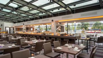 A modern cafe with multiple seating areas, a central counter with food display, and large windows allowing natural light.