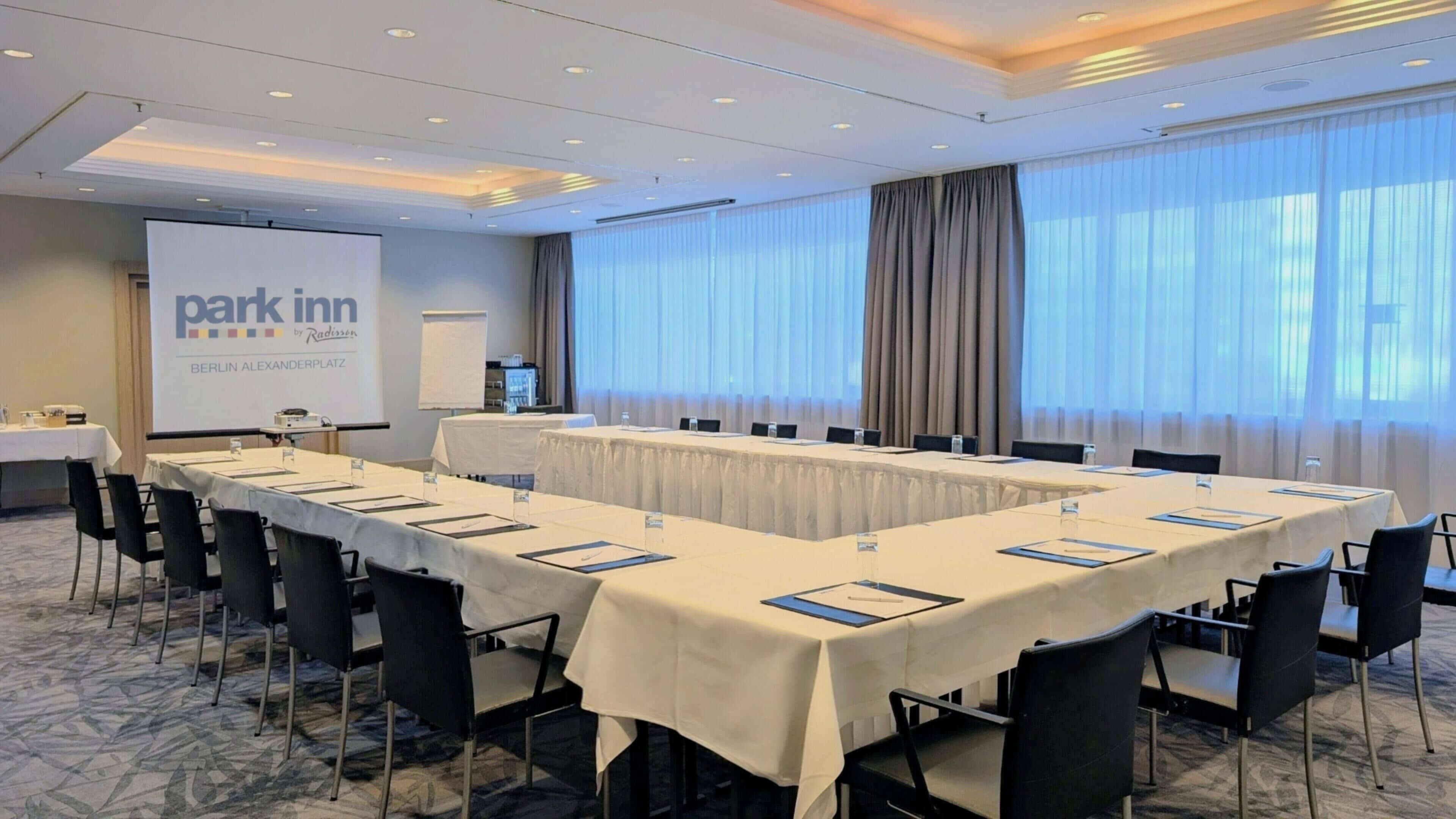 A conference room with a long table, chairs, a projector screen, and a presentation about Park Inn by Radisson Berlin Alexanderplatz.