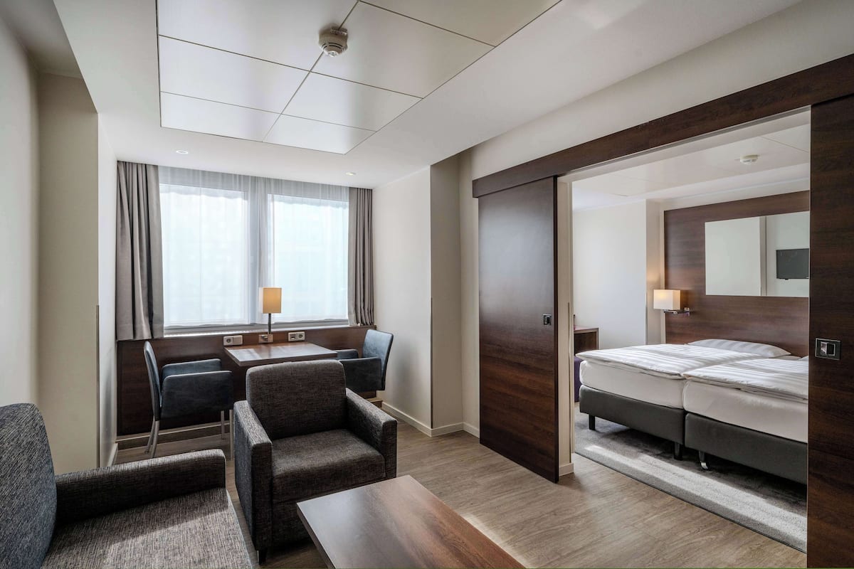 Habitación de hotel con cama, escritorio, dos sillas, ventana con cortinas y puerta corrediza que da a otra habitación.