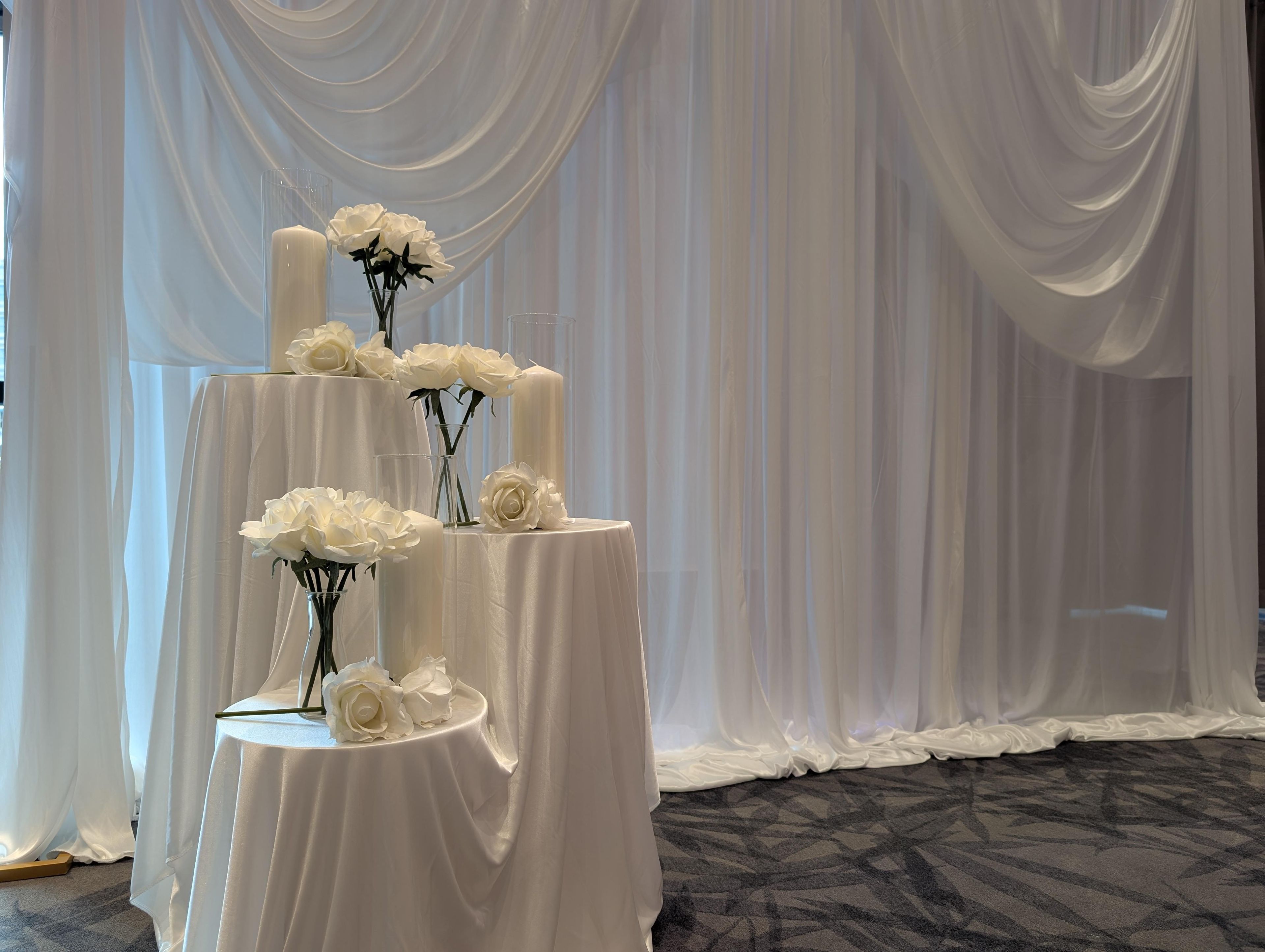Une table blanche à plusieurs niveaux, agrémentée de compositions florales et de bougies, le tout mis en valeur par un fond de draperies blanches.