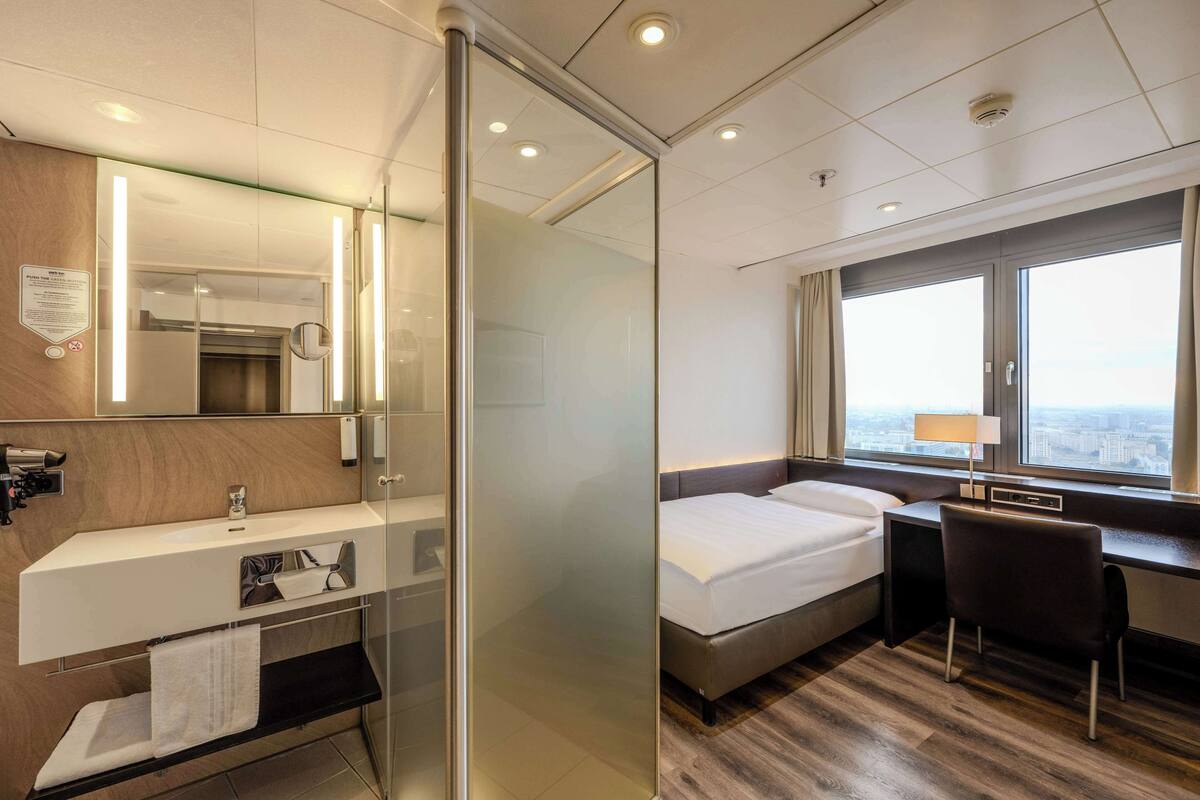 Habitación de hotel con separador de vidrio, cama, escritorio, silla, ventana con vista a la ciudad y baño con espejo y lavamanos.