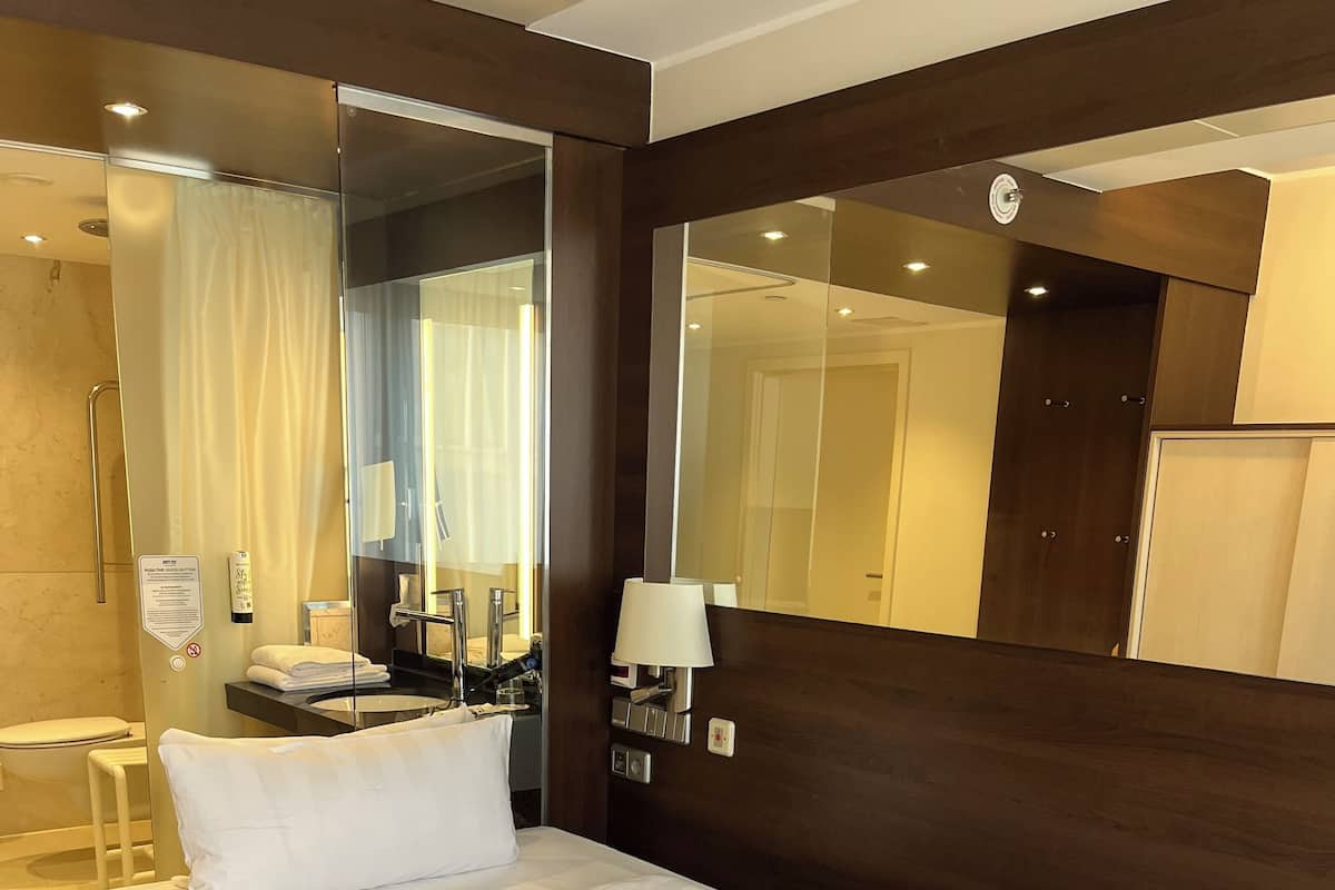 Una habitación de hotel con una cama grande, un baño con cabina de ducha de vidrio, un espejo y una mesita de noche con lámpara.