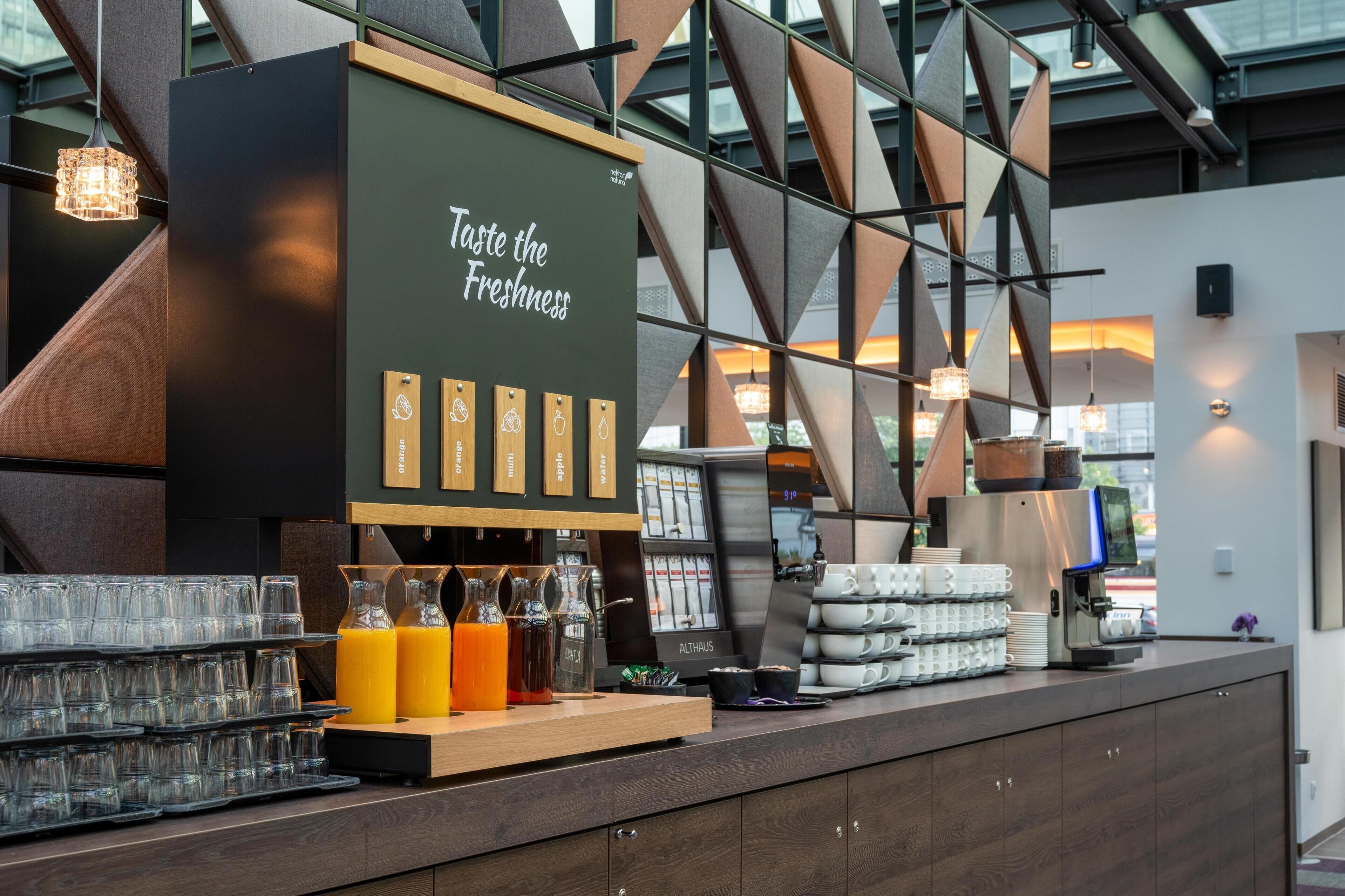A modern coffee station with a sign reading "Taste the Freshness," featuring a coffee machine, glassware, and juice dispensers.