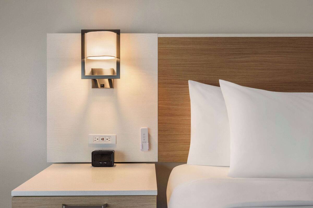 A hotel room with a bedside table, a wall-mounted lamp, a power outlet, and a control panel.