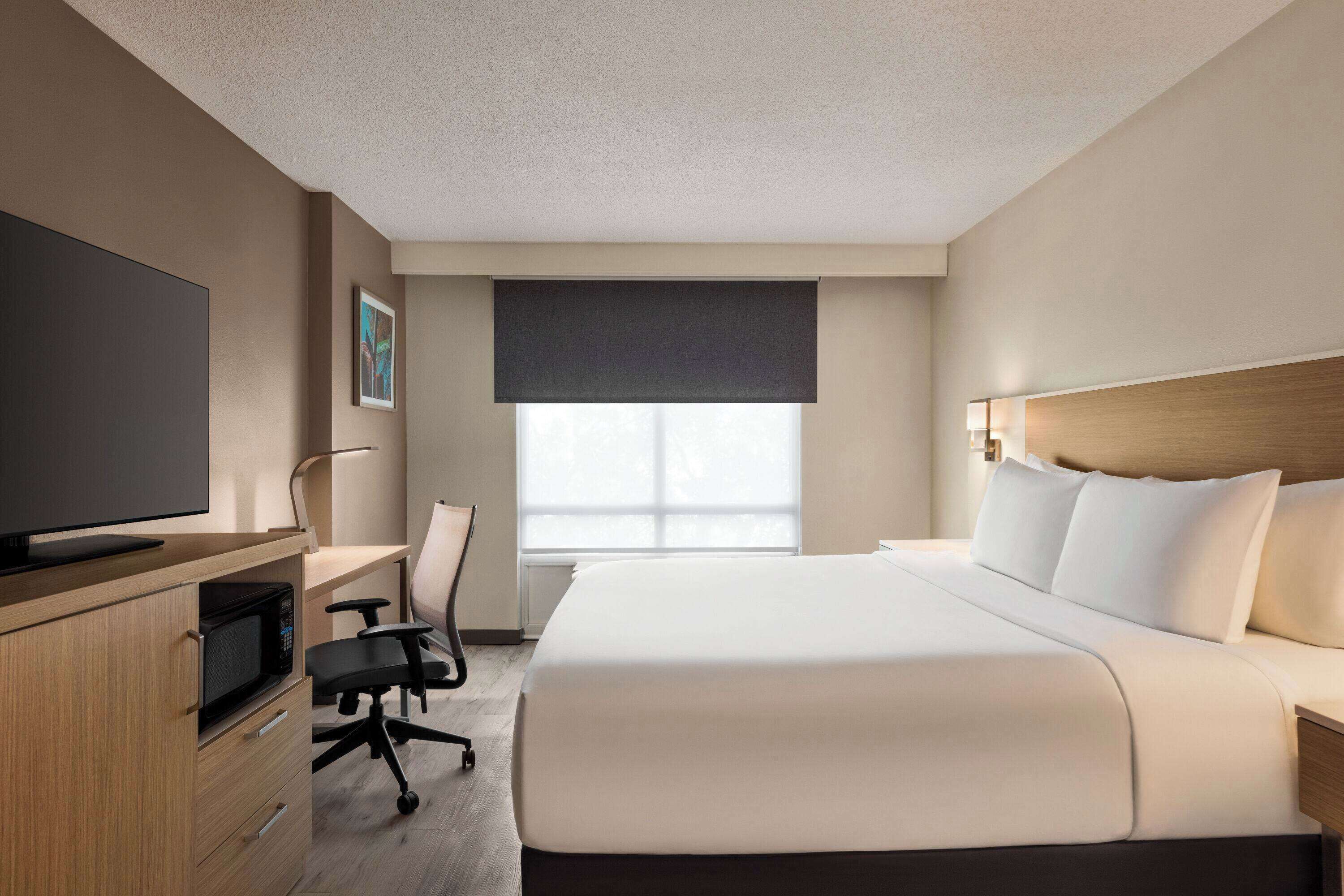 Una habitación de hotel con una cama, un escritorio con computadora, televisor y ventana con persianas.