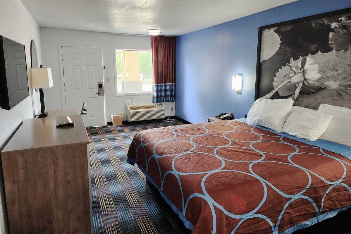 Habitación de hotel con cama, escritorio con lámpara, calefactor, teléfono, ventana con cortinas y un cuadro floral en la pared.