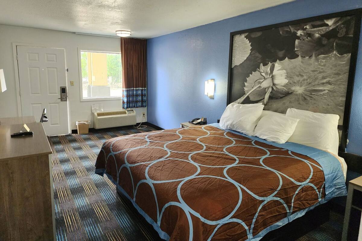 Habitación de hotel con cama, escritorio, ventana con cortinas, radiador y una lámpara fijada a la pared.